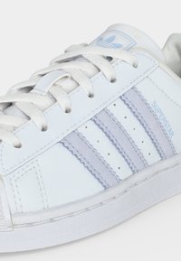 Λευκό sneaker Adidas Superstar με ανοιχτό γκρι ρίγες, λευκά κορδόνια και μπλε λογότυπο της μάρκας στη γλώσσα και στην πλευρά.