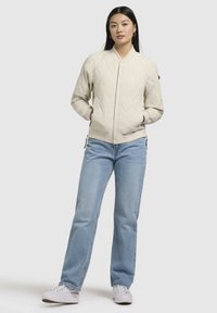 Vadderad beige bomberjacka med ribbad krage, framknäppning med tryckknappar och sidofickor, tillsammans med ljusblå jeans och vita sneakers.