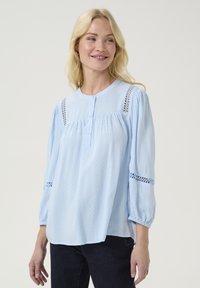 Gaiši zilas blūze ar noapaļotu apkaklīti, pogu aizdari, teksturētu audumu un mežģīņu apdari uz pleciem un piedurknēm. Brīvs piegriezums.