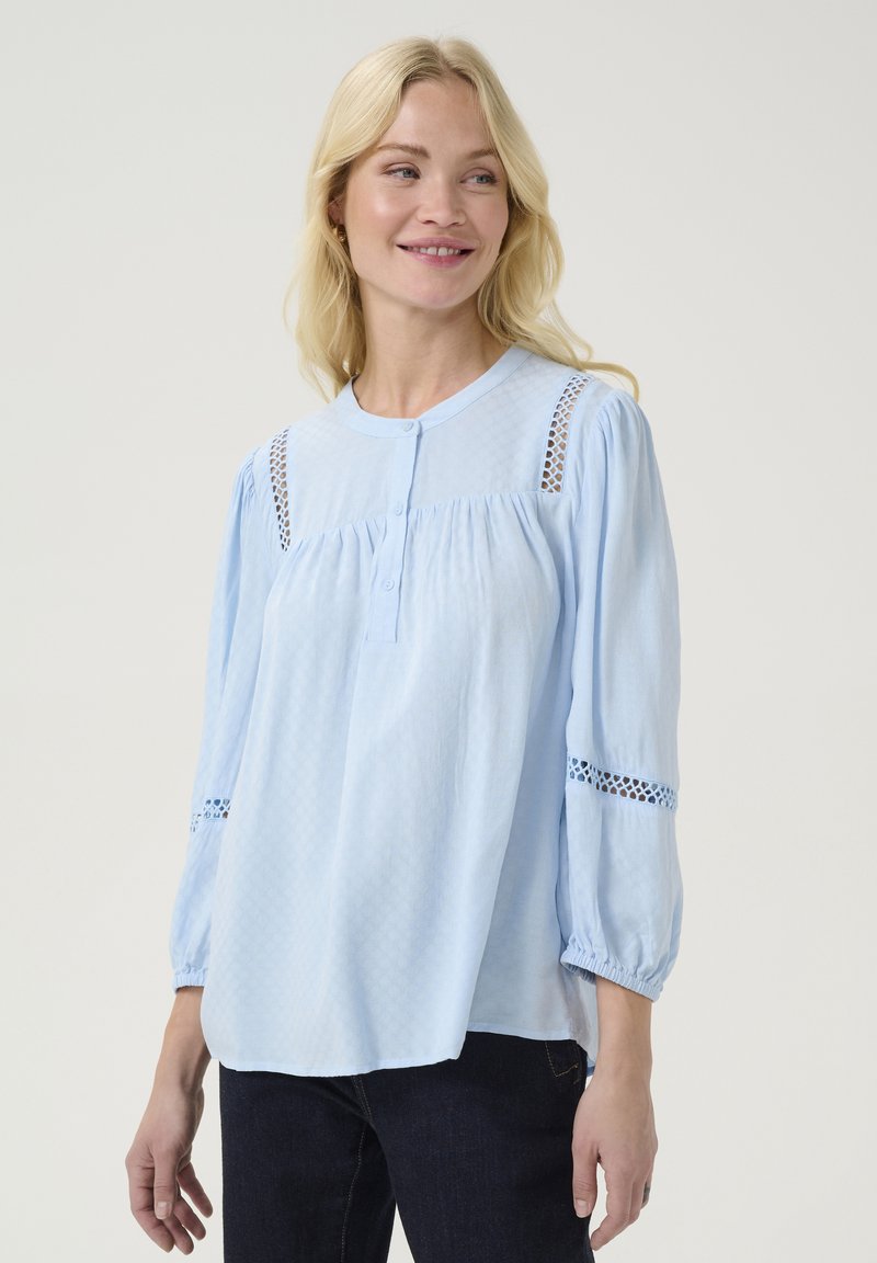 Gaiši zilas blūze ar noapaļotu apkaklīti, pogu aizdari, teksturētu audumu un mežģīņu apdari uz pleciem un piedurknēm. Brīvs piegriezums.