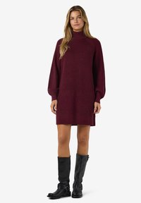 Vestido de malha bordeaux com gola alta, mangas longas e oversized, e bordas caneladas, combinado com botas pretas até aos joelhos.
