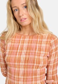 Chemise en tissu doux et texturé dans des teintes orange chaud avec des rayures rose et crème. Elle a un col bateau et des manches longues.