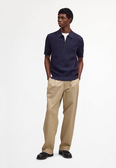 Mies seisoo taskut kädessä, yllään tummansininen lyhythihainen vetoketjuinen kauluspaita, beiget housut ja mustat juhlakengät, valkoista taustaa vasten.