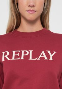 Sweatshirt bordeaux avec un col côtelé et des manches longues, présentant le mot "REPLAY" en grandes lettres majuscules blanches sur le devant.