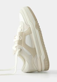 Beige och vit sneaker med tjock sula, snörning framtill, mesh och perforerade detaljer, visad stående på sin sula.