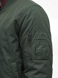 Gros plan d'une manche de veste bomber verte avec une poche zippée et des emplacements cousus pour stylos sur le haut du bras.