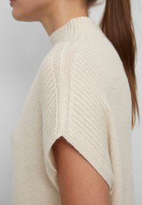 Pull léger beige avec un col montant, texture côtelée et manches courtes. Le tricot est doux et légèrement ample.