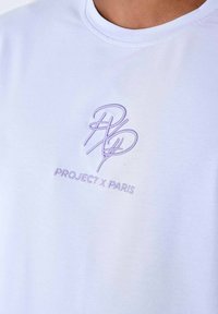 Camiseta blanca de tejido suave con logo bordado en púrpura "PXP" y texto "PROJECT X PARIS" en el pecho. Cuello redondo clásico.
