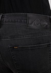 Svarta denimjeans med en läderlapp med "Lee"-logotypen, synlig sömmar och ett litet märkesetikett på bakfickan.