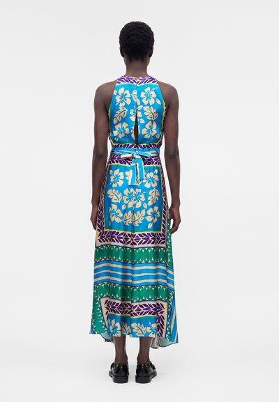 Γυναίκα φοράει αμάνικο φόρεμα με δέσιμο στην πλάτη, με μπλε, πράσινα και μωβ λουλουδένια και γεωμετρικά μοτίβα, στέκεται σε απλό λευκό φόντο.