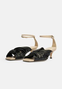 Czarne skórzane sandały na otwartym palcu z węzełkowym detalem na górze, beżowy pasek na kostkę, mały obcas i gładka, płaska podeszwa.