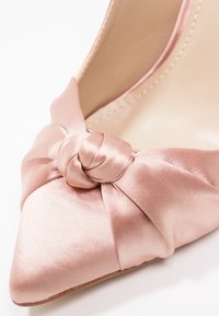 Escarpin en satin rose à talon haut, avec un bout pointu et un accent noué près de l'empeigne. Texture lisse avec un léger éclat.