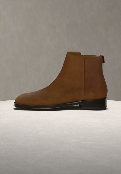 Bottine en daim marron avec un talon bas empilé et une fermeture éclair latérale, exposée sur une surface en pierre claire sur un fond neutre.