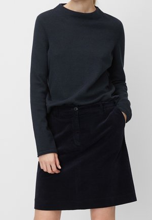 Vrouw draagt een donkerblauw langärmelig shirt dat in een bijpassende donkerblauwe ribfluwelen rok is gestopt, met één hand in de zak en de andere ontspannen.