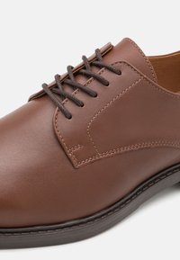 Scarpa di pelle marrone con texture liscia e dettagli cuciti. Presenta lacci marroni scuri e una punta arrotondata. La suola è in gomma nera.