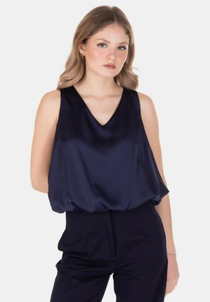 Giovane donna con capelli biondi fino alle spalle, indossa una blusa in raso blu navy senza maniche e pantaloni scuri abbinati su sfondo bianco.