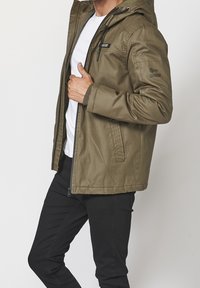 Mężczyzna w oliwkowej kurtce z kapturem, nałożonej na białą koszulę i czarne spodnie, trzymający rękę na suwaku kurtki.