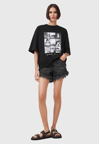 Camiseta negra de gran tamaño con diseño de collage fotográfico, combinada con pantalones cortos de mezclilla negra desgastados con flecos en el dobladillo y sandalias planas.