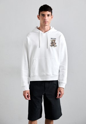 Witte hoodie met een berenafbeelding en de tekst "Moschino", voorzien van een voorzak en een trekkoord in de capuchon. Gecombineerd met zwarte shorts.