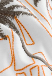 Tissu blanc avec broderie texturée orange et grise formant des motifs géométriques et linéaires abstraits.