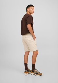 T-shirt marron associé à un short beige clair. Le short est doté de poches latérales et arrière. L'ensemble est complété par des baskets marron et gris.