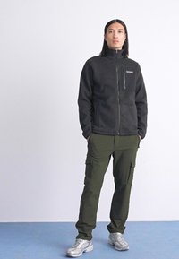Persoon die met handen in de zakken staat, gekleed in een zwarte fleece zipjack, olijfgroene cargobroek en zilveren sneakers op een blauwe vloer tegen een witte muur.