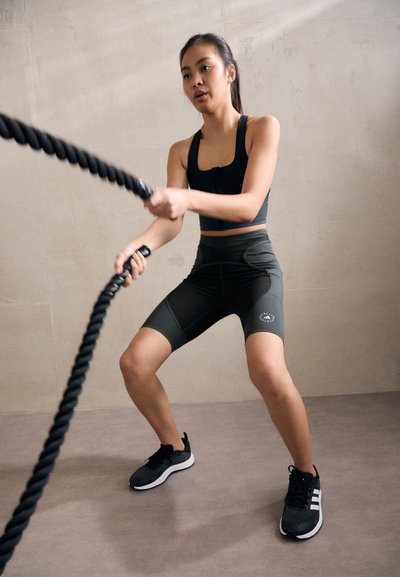 Black sports bra and dark green high-waisted shorts, with a logo. The person is using thick black battle ropes in a gym setting.
