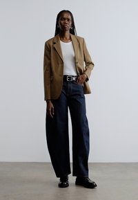 Brun skræddersyet blazer over hvid tætsiddende tanktop, højttaljede mørkeblå vidde ben jeans og sorte lædersko. Ensfarvet baggrund.