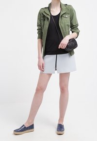 Grön lättviktsjacka, svart linne, ljusblå kjol och marinblå espadrillos. Svart crossbody-väska, dragkedjedetalj på kjolen.