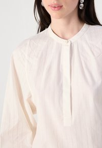 Primo piano del collo di una persona che indossa una camicia leggera a righe con pattina dei bottoni, che mette in evidenza le maniche lunghe e delle pieghe sottili.