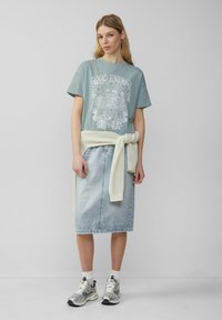Lichtblauw T-shirt met witte bloemenprints, gecombineerd met een lichte denim rok en een crèmekleurige gebreide trui om de taille geknoopt. Zilveren sneakers.
