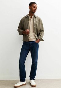 Helles olivgrünes Shirt-Jackett, beige Polohemd, dunkelblaue Jeans und weiße Sneaker mit braunen Sohlen. Einfaches Design und klassischer Schnitt.