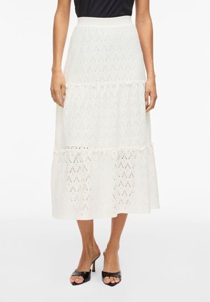 Žena oblečená v bielej midi sukni s ozdobnými dierkami a volánmi, v kombinácii s čiernymi otvorenými sandálmi na podpätku a čiernym topom.