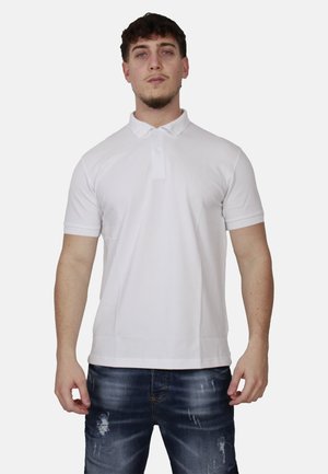 Giovane uomo con indosso una polo bianca a maniche corte e jeans blu scuro consumati, in piedi contro uno sfondo chiaro.