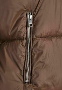 Veste rembourrée marron avec une fermeture éclair silver visible. La texture est lisse avec un motif matelassé, présentant des sections piquées.