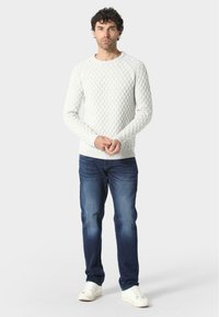 Witte gebreide trui met een gestructureerd ruitpatroon, geribbelde manchetten en zoom, gecombineerd met donkerblauwe denim jeans en witte sneakers.