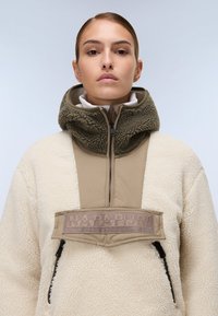 Μπεζ φλιτζάνι μπουφάν με ψηλό γιακά, με κουκούλα επενδεδυμένη με σκούρο πράσινο ψεύτικο γούνινο υλικό και με λογότυπο πάνω στο στήθος. Τσέπη με φερμουάρ στο πλάι.