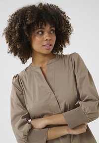 Kergelt pruun kaelusega pluus, millel on puffse varrukad ja nööpdetail kaela ääres; sile tekstuur ja mansetiga varrukad.