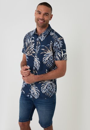 Smilende mand iført en kortærmet navyblå tropisk mønstret skjorte og cowboyshorts, stående mod en ensfarvet lys baggrund.