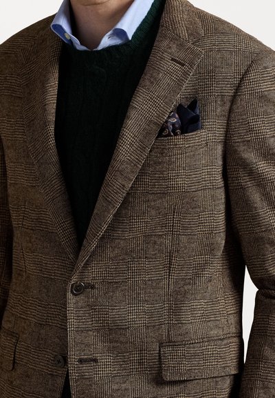 Blazer marrón a cuadros con un acabado texturizado, dos botones y un pañuelo de bolsillo. Superpuesto sobre un suéter de punto de trenzas verde oscuro.