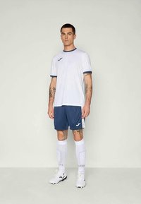 Bílý sportovní dres s námořnickými akcenty, doplněný námořnickými šortkami. Bílé ponožky a černé kopačky dotvářejí outfitu. Viditelné logo sportovní značky.