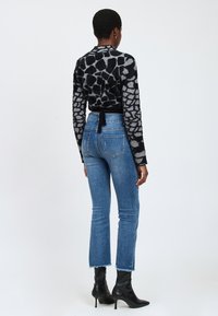Donna in piedi, che indossa un maglione a fantasia giraffa nero e grigio, jeans blu a vita alta con orli sfrangiati e stivaletti neri con tacco a punta.