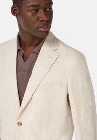 Blazer de lino beige con cierre de un solo botón, solapas de pico y bolsillo en el pecho. Lleva puesto una camiseta polo marrón.