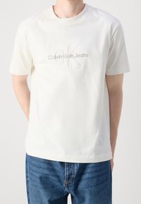 Bílá bavlněná trička s krátkým rukávem, s texturovaným logem "CK" a výšivkou "Calvin Klein Jeans" v světle hnědé barvě.