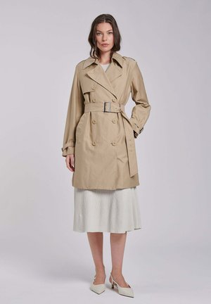 Donna che indossa un trench beige con cintura sopra un vestito midi bianco e scarpe bianche con tacco basso, in piedi su uno sfondo semplice.