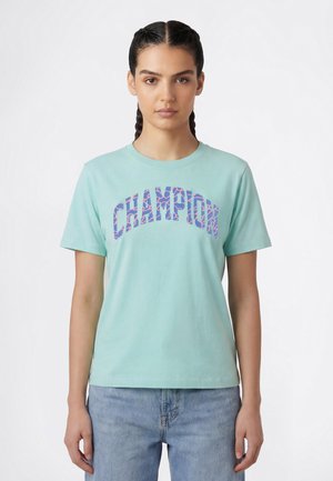 Kobieta z zaplecionymi włosami, ubrana w jasnoniebieską koszulkę z kolorowym napisem „CHAMPION” oraz jasnoniebieskie dżinsy, stojąca na tle jednolitego białego tła.