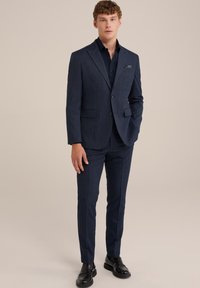 Costume bleu marine en tissu texturé, composé d'une veste à deux boutons avec revers et d'un pantalon slim, associé à des chaussures noires.