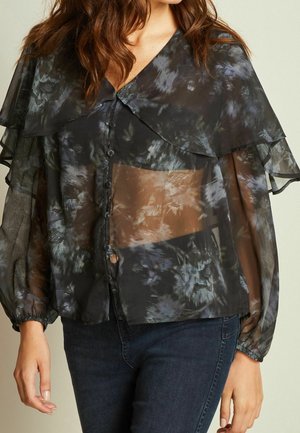 Vrouw draagt een doorschijnende zwarte blouse met bloemmotieven, een oversized kraag, lange opgeblazen mouwen en een knoopsluiting aan de voorkant, gecombineerd met donkere spijkerbroek.