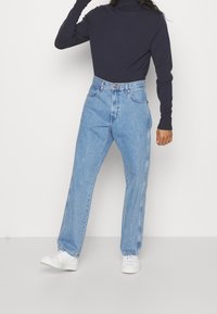 Hellblaue hochgeschnittene Jeans mit geradem Bein, sanften Färbungseffekten, Gürtelschlaufen und klassischem Fünf-Taschen-Design, kombiniert mit einem schwarzen Rollkragenpullover und weißen Sneakern.