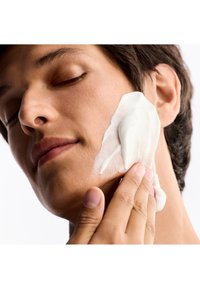 Facial cleanser application on skin; creamy white texture spreading across the cheek with a hand. Smooth, bare skin backdrop.